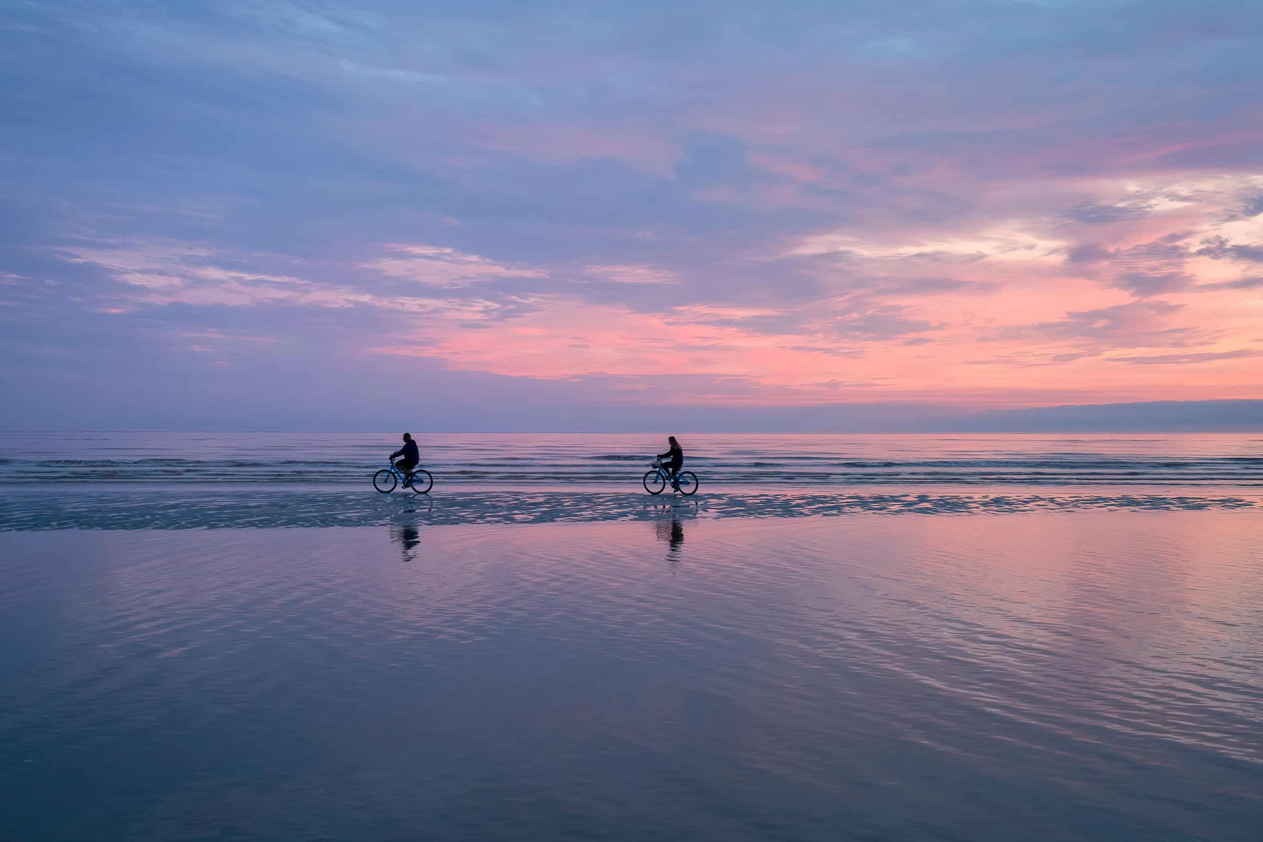 Bicycles riding along the beach at sunset, with the purple and pink sky reflected in the water.