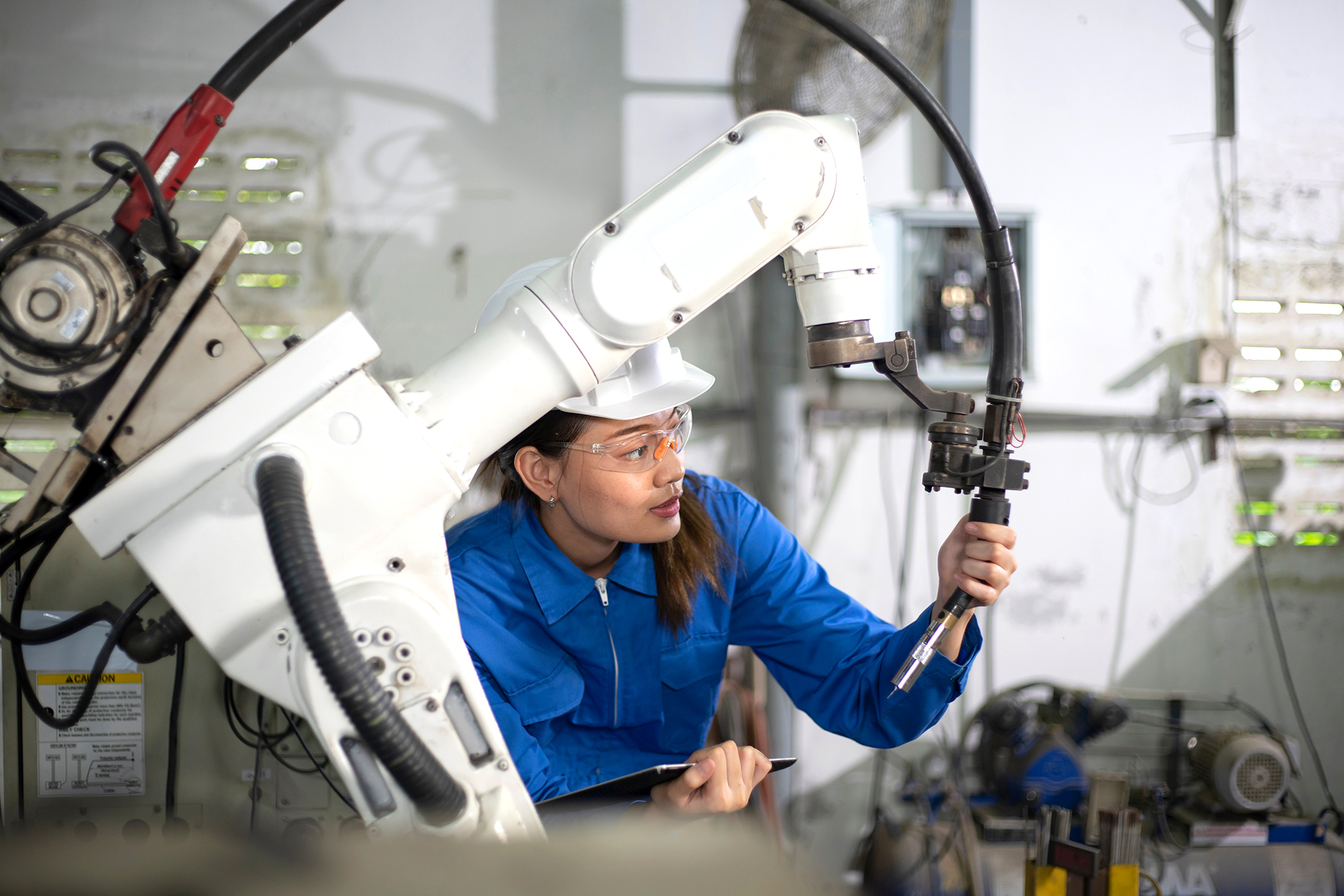 EV worker in a blue mechanic's suit and hardhat working with a robot arm