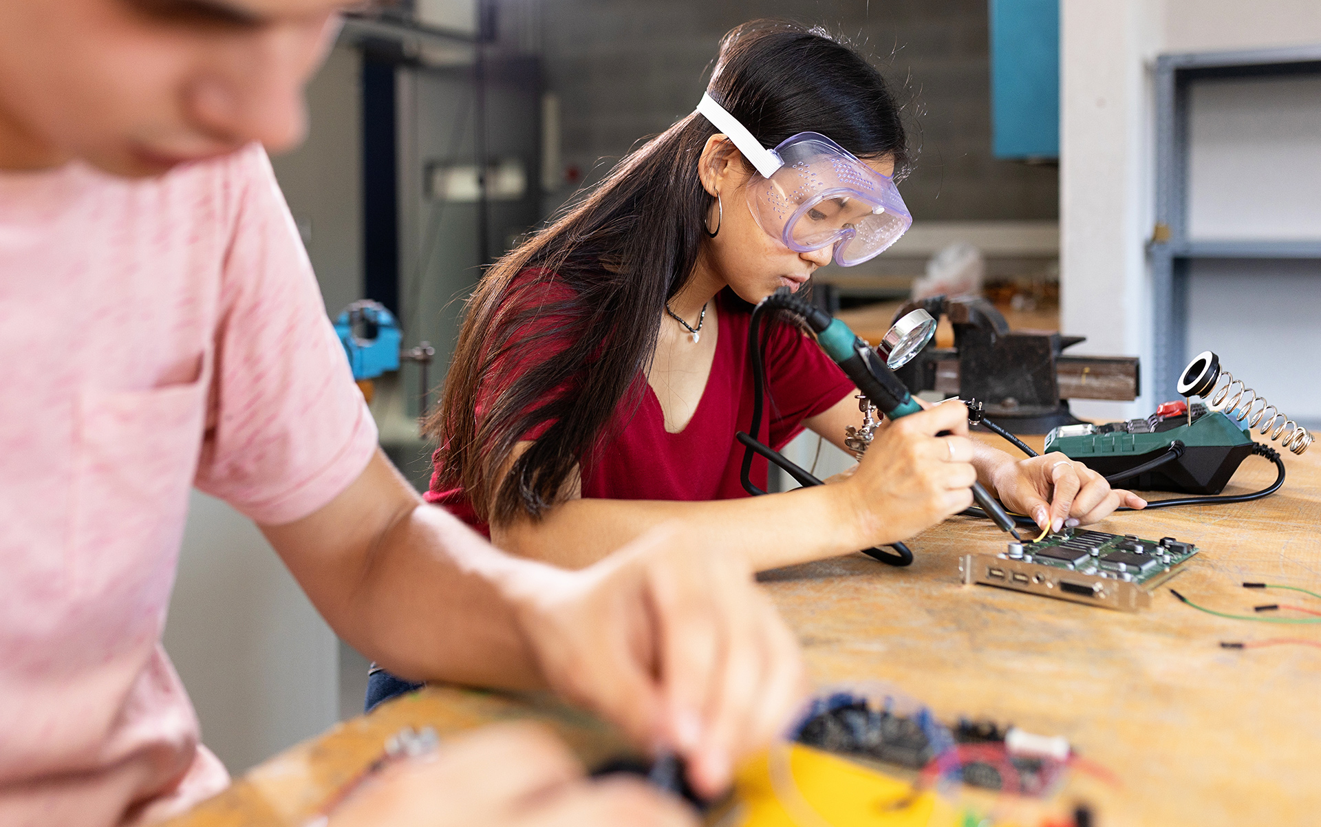 Workers assembling electronics at a workbench while wearing protective eyewear.