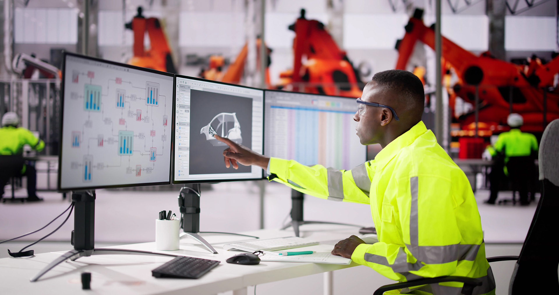 A man in a bright safety shirt working at a desk with three large monitors. Big orange assembly robots are in the background.